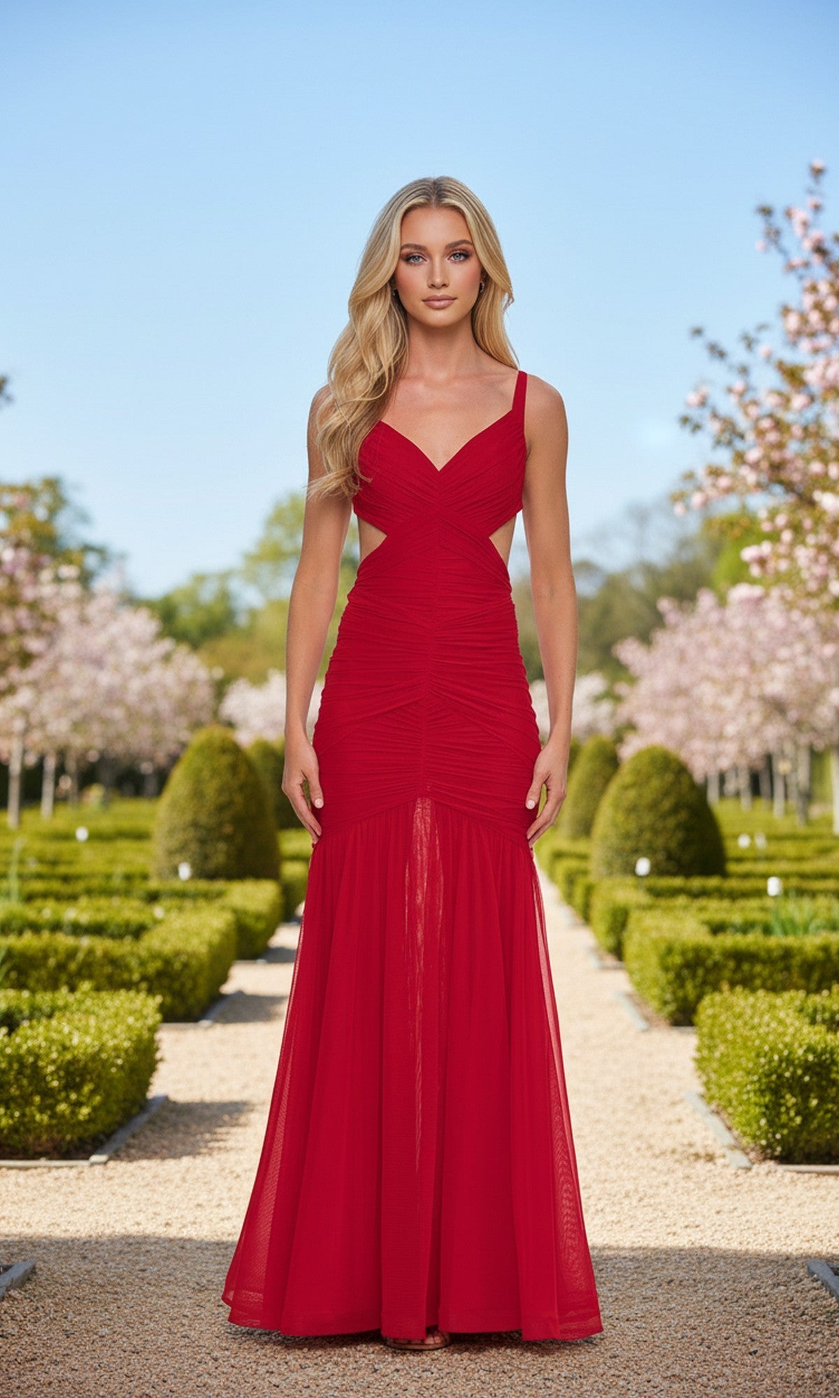 A woman in the La Femme 33626 long red prom dress stands on a gravel path in a manicured garden with blooming trees and trimmed bushes.